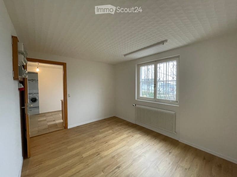 Empty room with wooden flooring, white walls, and a large window. A radiator is visible under the window, and a door leads to another room with a washing machine.