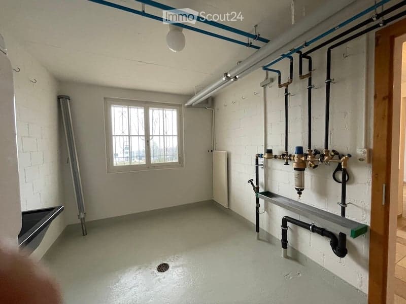 Empty utility room with white brick walls, concrete floor, window, exposed pipes, and a drain in the center.