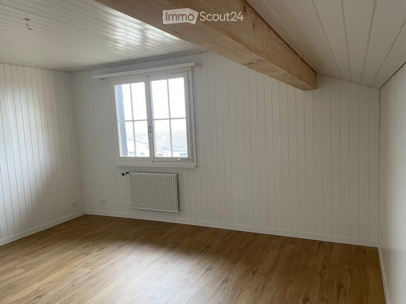 Empty room with wooden flooring, white paneled walls, a single window with white frame, and a radiator below the window.