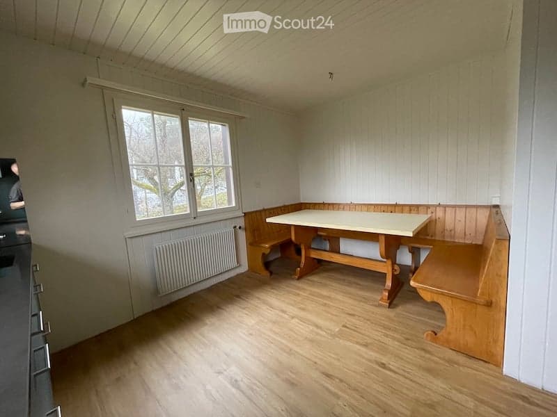 Bright, wooden-floored room with a large window, a built-in wooden bench seating, and a central wooden table.