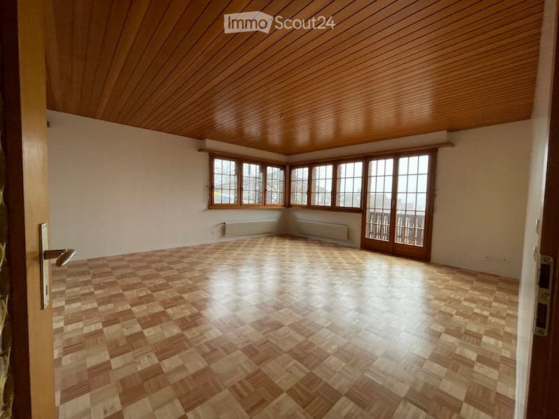 Empty room with wooden parquet flooring, wooden paneled ceiling, large windows, and a sliding glass door leading to a balcony.