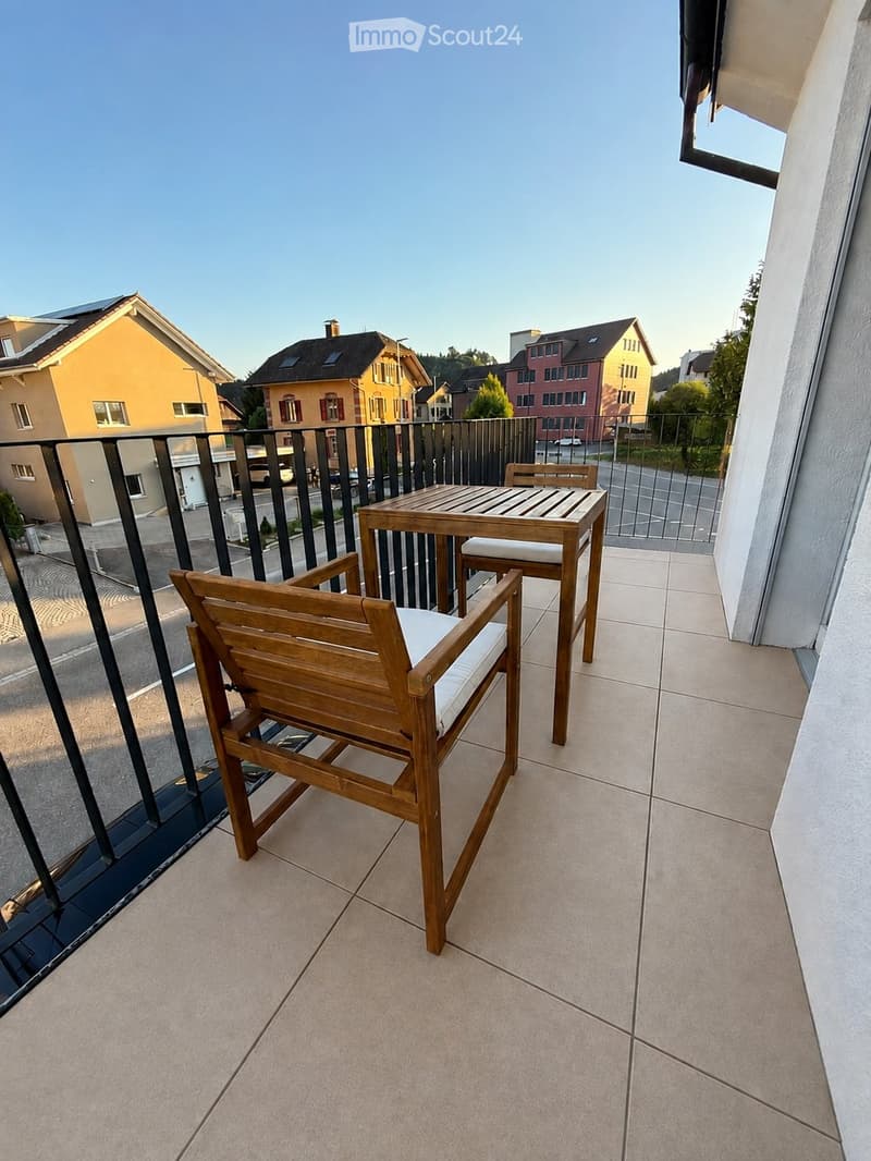 Balcony with tiled floor, wooden table, and two wooden chairs with white cushions, overlooking a residential area.