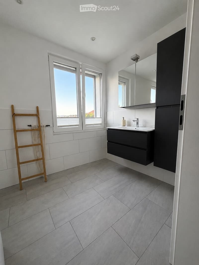 Modern bathroom with white tiled walls, grey tiled floor, large window, black vanity cabinet, mirror, and a wooden drying rack.