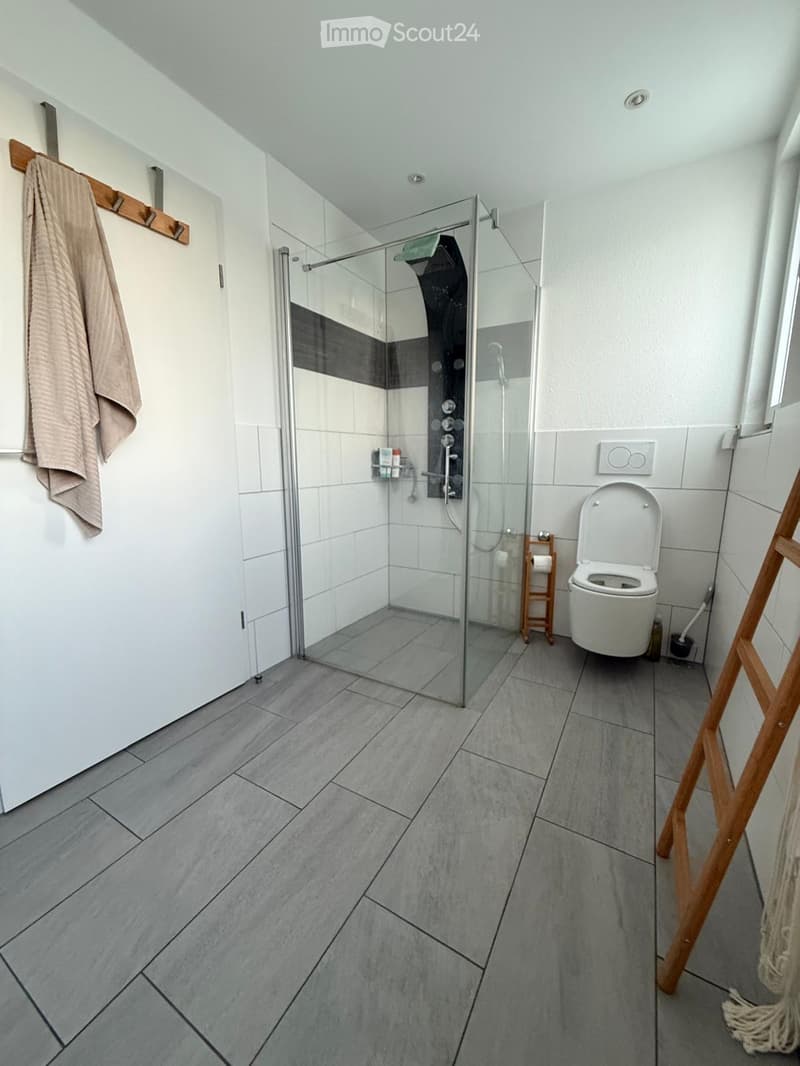 Modern bathroom with grey tiled floor, white walls, glass-enclosed shower, toilet, towel on a wooden rack, and a small wooden shelf holding toiletries.