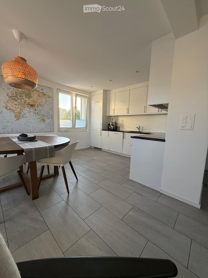 Modern kitchen with white cabinets, a round dining table with chairs, and a hanging woven lamp. The floor is tiled, and there is a world map on the wall.