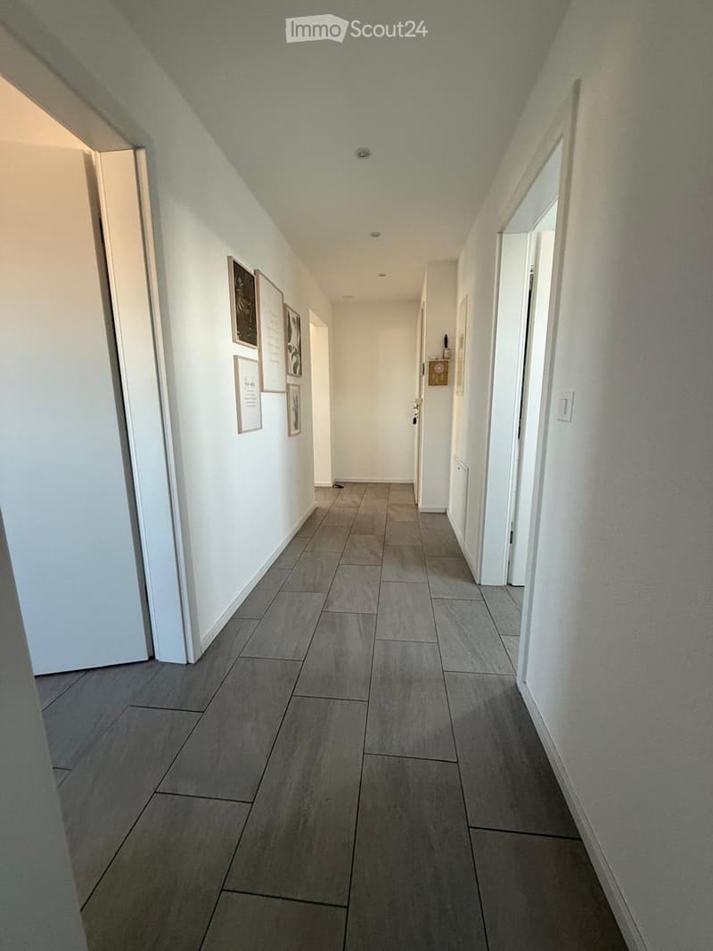 Clean, modern hallway with gray tiled flooring, white walls, and several doors leading to different rooms. A small table with a candle is visible on the right side.