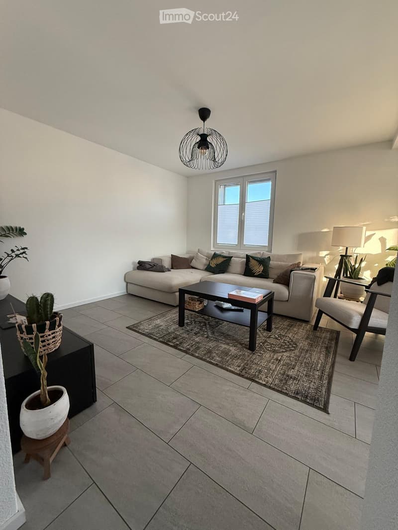 Modern living room with a grey sofa, coffee table, armchair, and decorative plants. Features a tiled floor, large window, and contemporary lighting.
