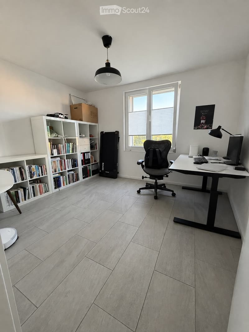 Modern office space with light grey tiled flooring, white walls, large windows, a black office chair, a white desk with a laptop, keyboard, mouse, and a lamp, a bookshelf filled with books, and a small exercise bike in the corner.
