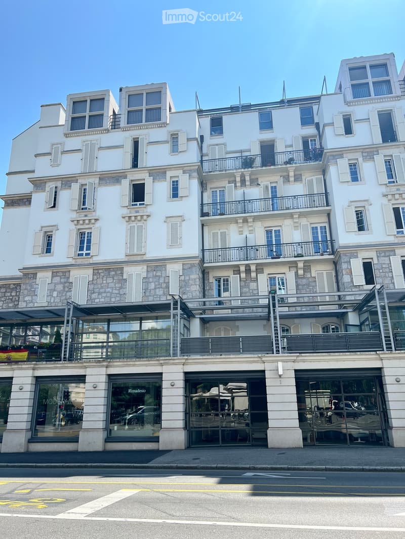 A multi-story white building with stone accents, multiple windows, balconies, and a modern storefront with glass doors and signage.