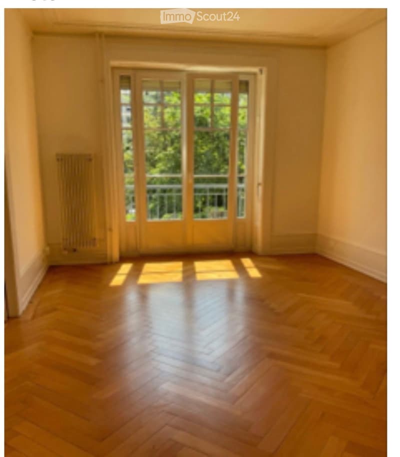Empty room with wooden flooring and large glass doors leading to a balcony. The walls are painted in a light color, and the room is well-lit with natural light.