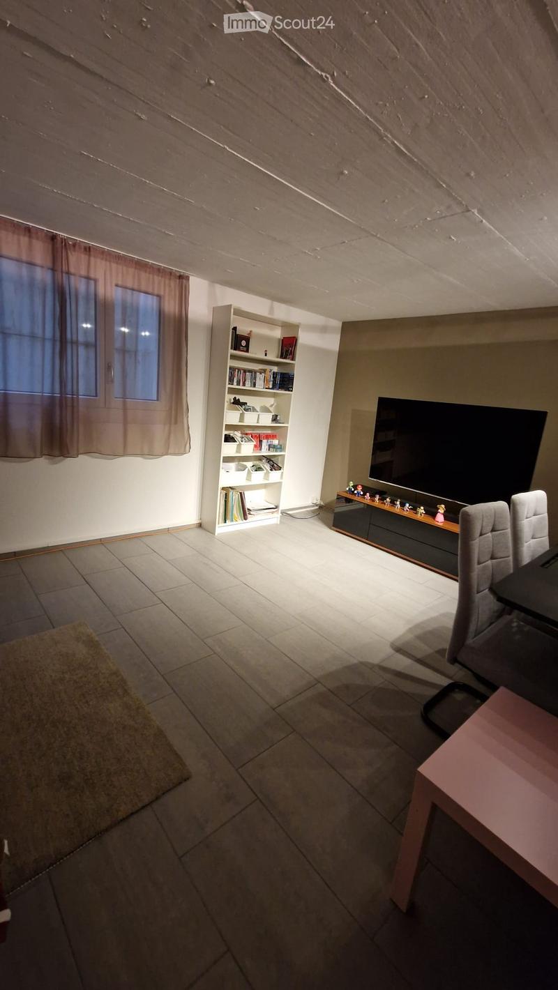 A modern living room with tiled flooring, a large flat-screen TV on a wooden stand, a bookshelf filled with books and decorative items, and a small table with chairs.