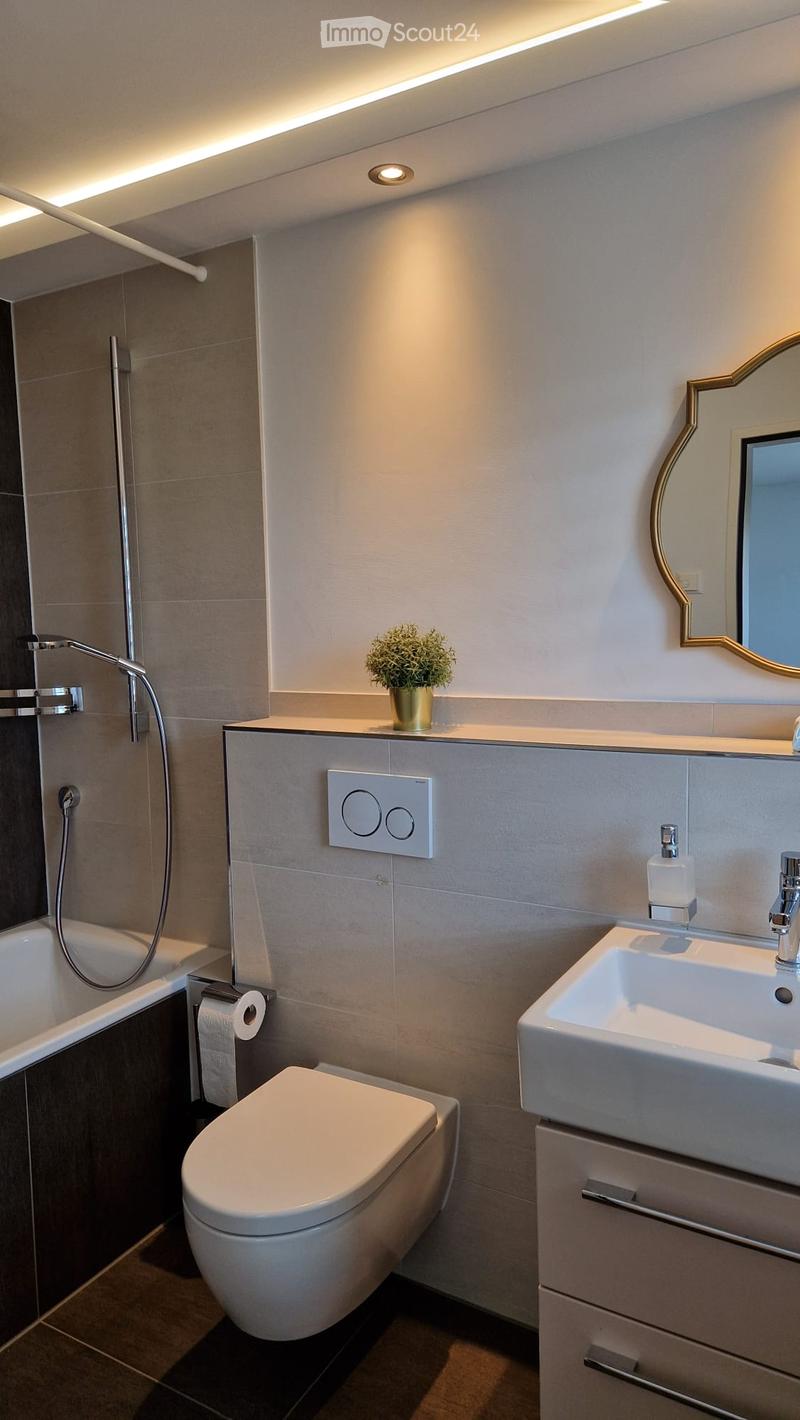 Modern bathroom with a white toilet, sink, bathtub, and a decorative mirror. Features a sleek design with a potted plant on the counter and a showerhead visible.