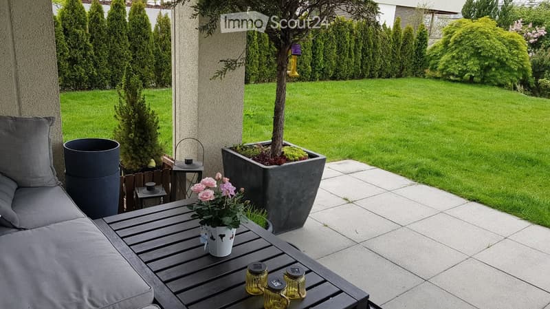 A well-maintained garden with a patio area featuring a wooden table, grey sofa, potted plants, and a small tree in a large planter. The lawn is neatly trimmed, and the area is surrounded by tall, manicured evergreen trees.