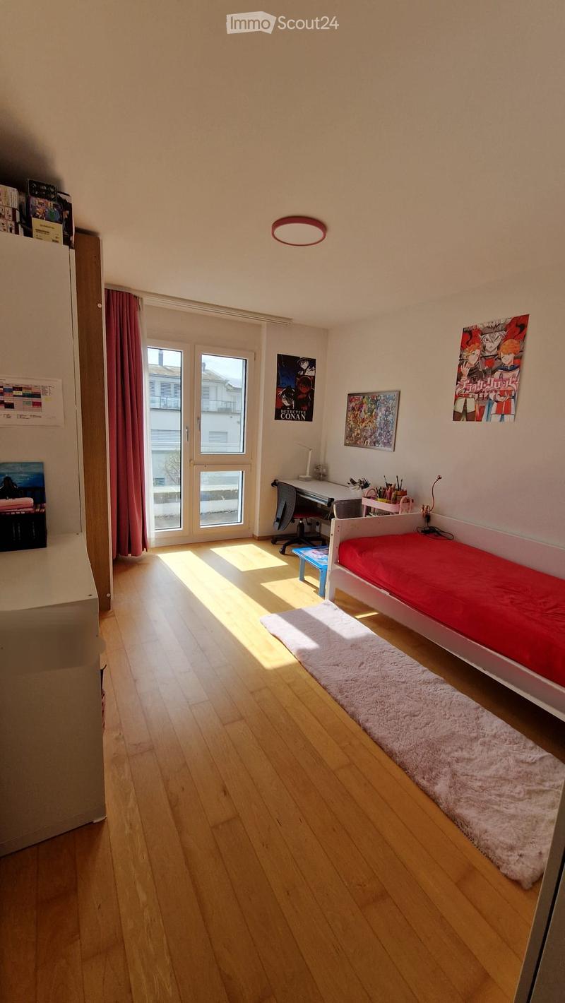 Bright bedroom with wooden flooring, a single bed with red bedding, a desk with a chair, and various posters on the walls. Natural light streams in through large windows with red curtains.