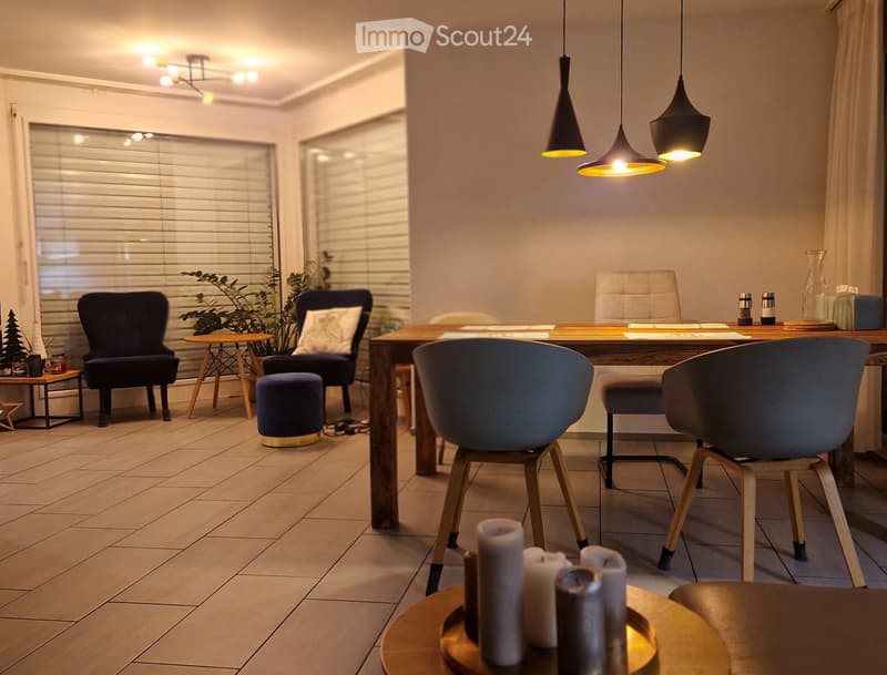 Modern living room with a mix of seating areas, including a sofa, chairs, and a small table. There is a large wooden table with chairs, a small round table with candles, and decorative items on a side table. The room features pendant lights and large wind