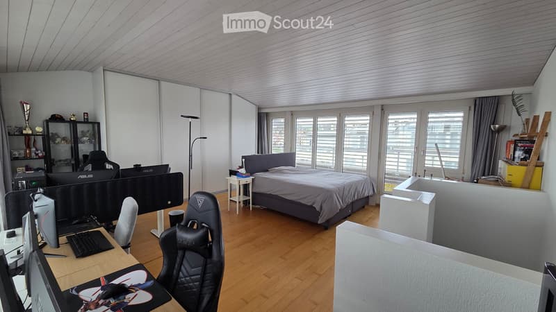 A multi-functional room with a bed, desk setup, and gaming area. Features a wooden floor, large windows, and modern furniture.