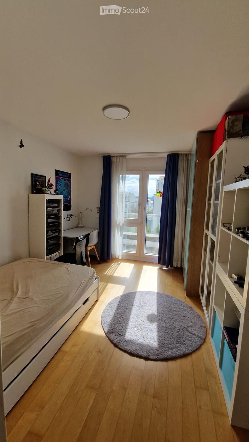 Cozy bedroom with a single bed, a round rug, a desk with a chair, and shelves. The room has wooden flooring and large windows with curtains leading to a balcony.