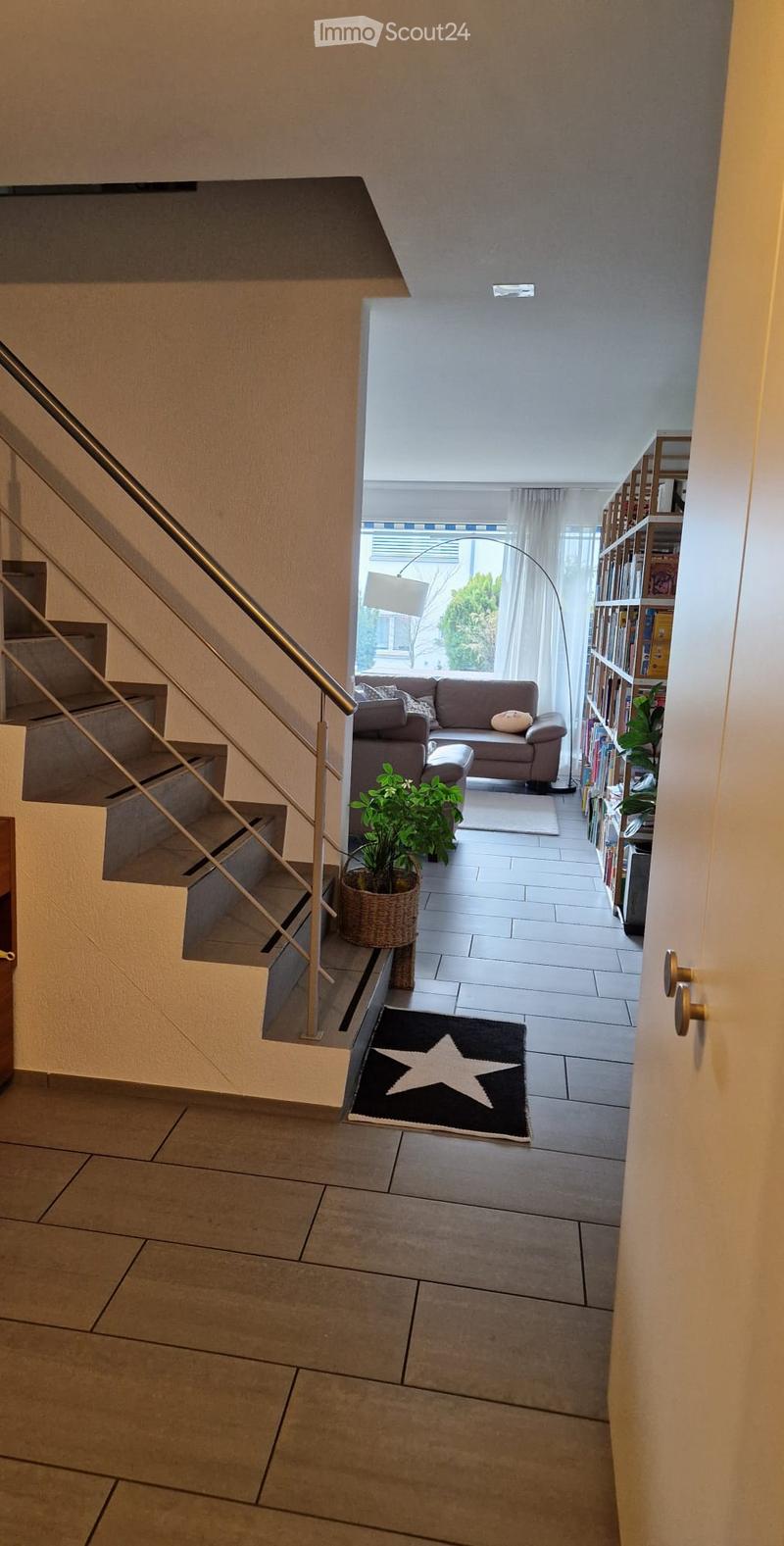 Modern hallway with tiled flooring, staircase with metal railing, and a view into a living area with a sofa, bookshelf, and plants.