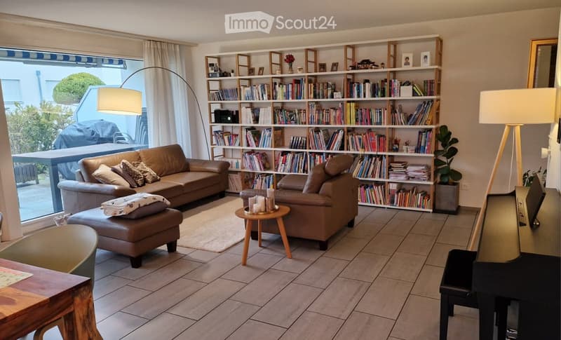 Living room with tiled floor, large window, bookshelf filled with books, leather sofa, ottoman, coffee table, floor lamp, and table lamp.