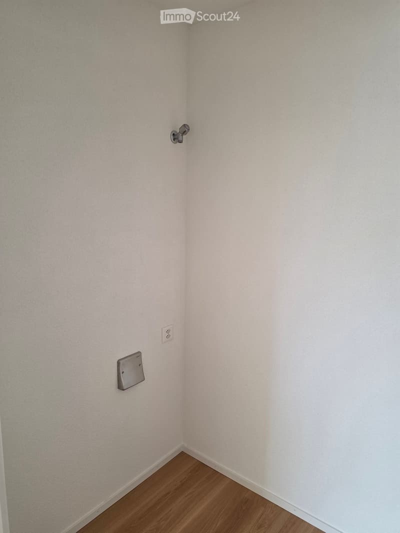 Empty room with light-colored walls and wooden flooring, featuring a light switch, an electrical outlet, and a small wall-mounted device.