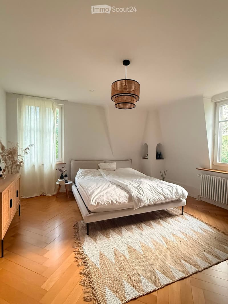 Bright bedroom with wooden flooring, a large bed with white bedding, a nightstand, a small table with a vase, and a hanging woven pendant light.