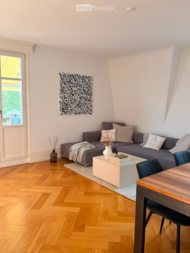 Modern living room with wooden herringbone flooring, grey sofa with decorative pillows, coffee table with books and vase, and a small dining area with a chair and table.