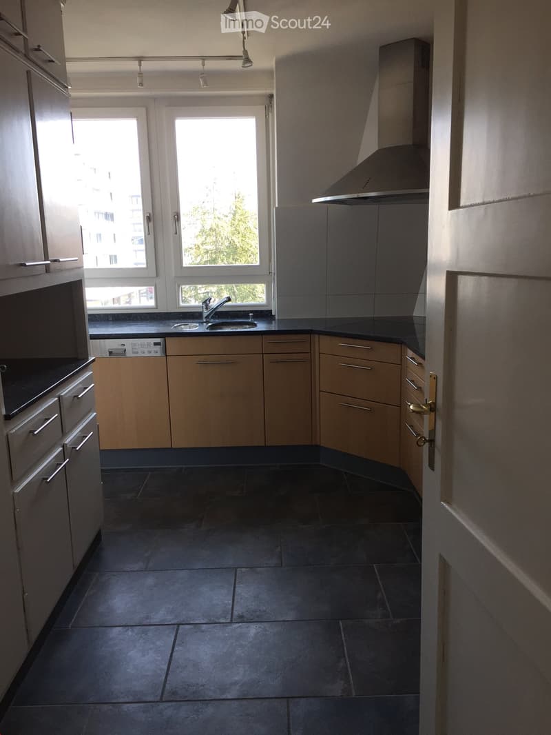 Modern kitchen with dark countertops, light wooden cabinets, and stainless steel appliances. Features a sink, faucet, and range hood.