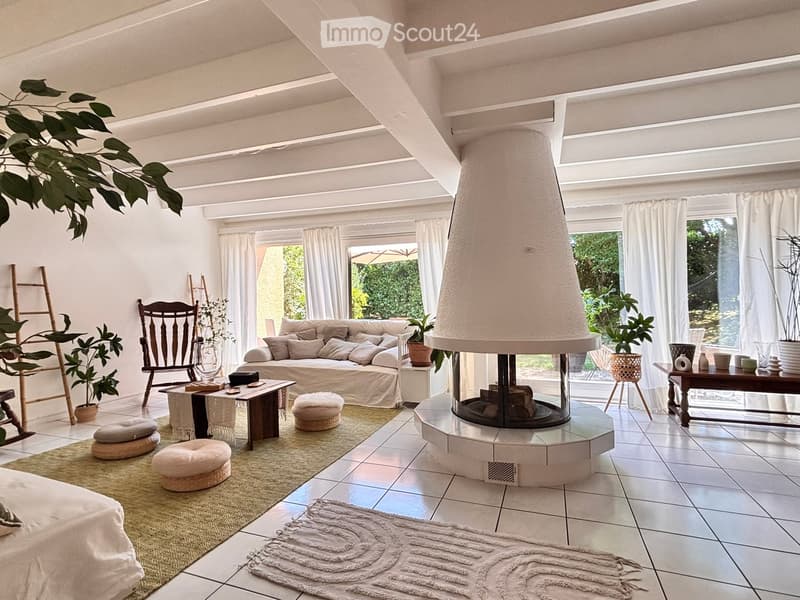 Bright living room with white walls, large windows, a central fireplace, a sofa, a coffee table, a wooden chair, and several plants.