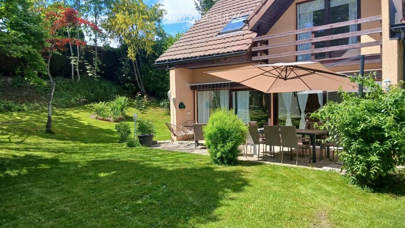 Garden with a well-maintained lawn, a patio area with a table and chairs, and an umbrella providing shade. The house has a balcony and is surrounded by trees and shrubs.