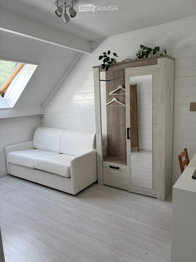 A modern, minimalist attic room with white wooden walls, a white leather sofa, a tall wardrobe with a mirror, and a small table with a chair.