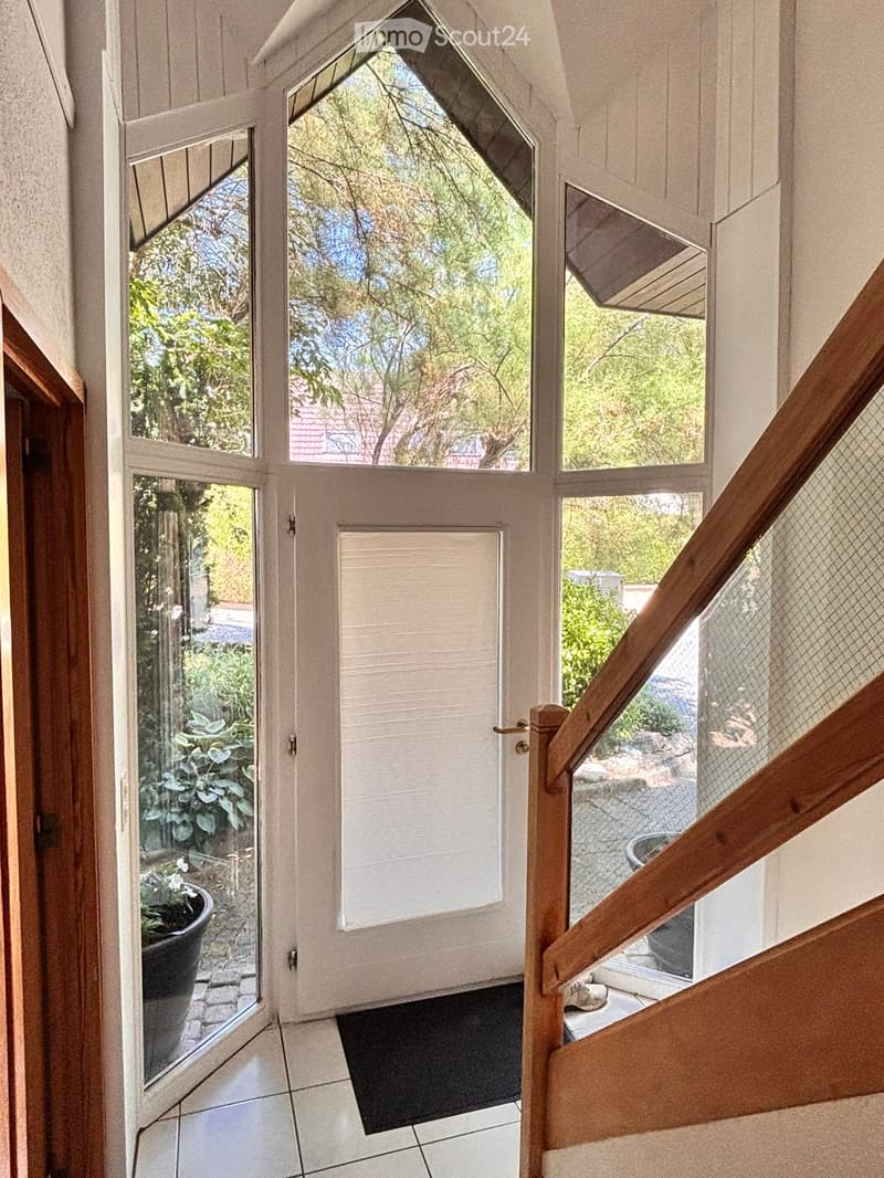 Bright hallway with large windows and a staircase leading upwards, featuring a tiled floor and a black doormat.