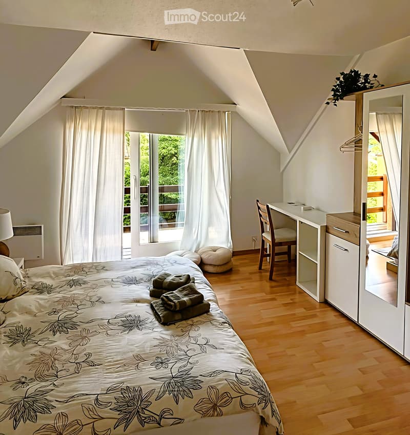 Cozy attic bedroom with wooden flooring, large window with white curtains, bed with floral duvet, mirror wardrobe, desk with chair, and two round stools.