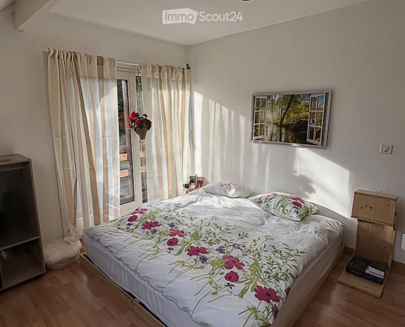 Cozy bedroom with a large bed, floral duvet, nightstand, mirror, and a picture frame on the wall. Light-colored curtains cover the windows, and a small dog bed is placed near the wardrobe.