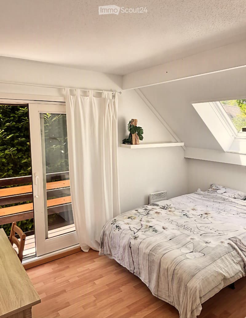 Cozy attic bedroom with wooden flooring, a double bed with floral bedding, a small table and chair near the window, and a shelf with a plant. The room features a skylight and a sliding glass door leading to a balcony.