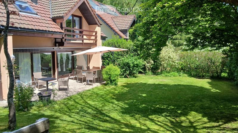 Lush green garden with a patio area featuring a table, chairs, and an umbrella, adjacent to a modern house with a balcony and large windows.