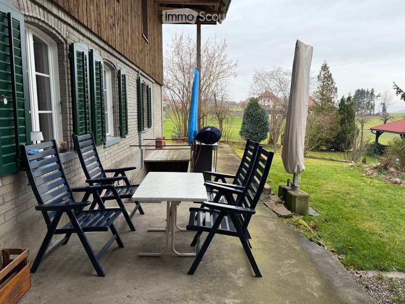Balcony with a table and two chairs, overlooking a grassy area with trees and a house in the background.