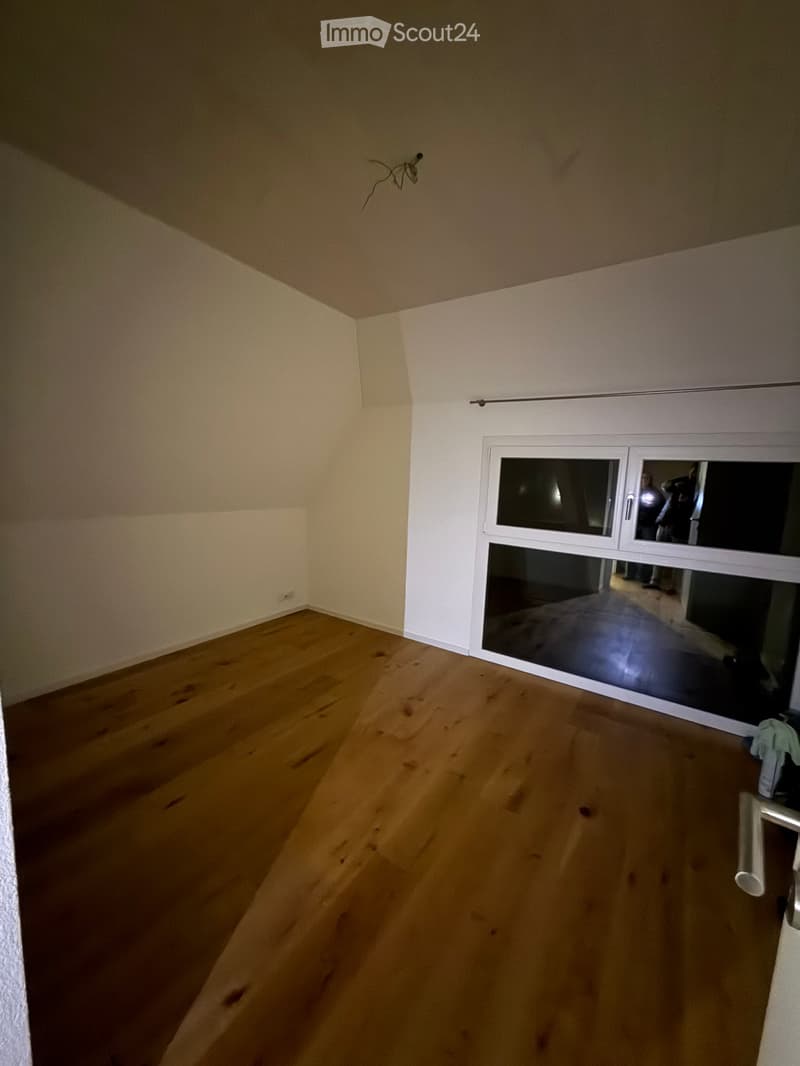 Empty room with wooden flooring and white walls, featuring a large window with a view into another room.