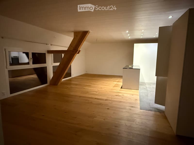Empty room with wooden flooring, white walls, and recessed ceiling lights. A small white counter is visible in the corner, and a large window with blinds is on the left side.