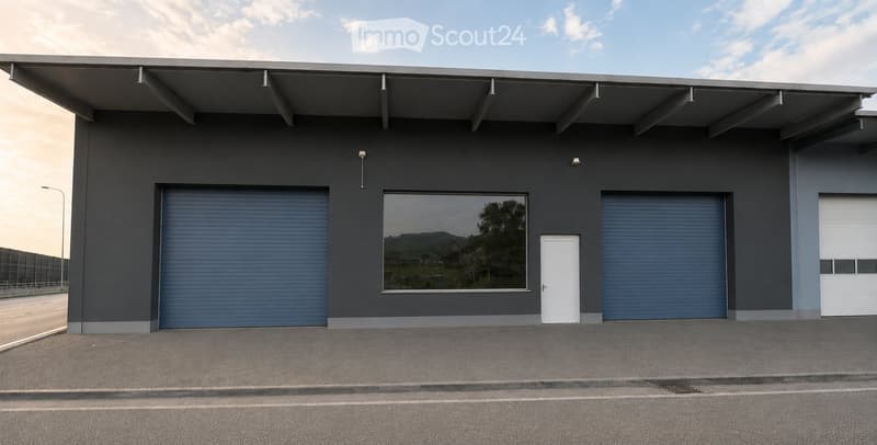 Modern industrial-style building with grey walls, large garage doors, and a single entrance door. Features a large window and a flat roof with extended overhang.