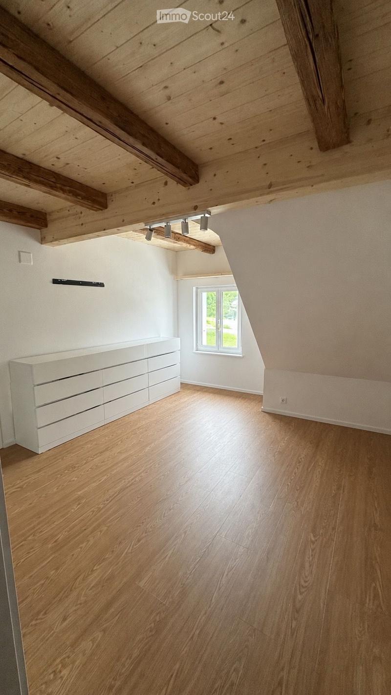 An empty attic room with wooden floors, white walls, exposed wooden beams, and a single window letting in natural light. The room features built-in white storage units along one wall and recessed lighting fixtures on the ceiling.