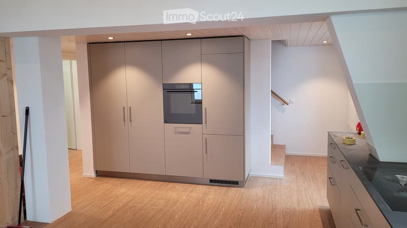 Modern kitchen with light wood flooring, built-in cabinets, and an oven. A small countertop with a stool is visible, and a broom leans against a pillar.