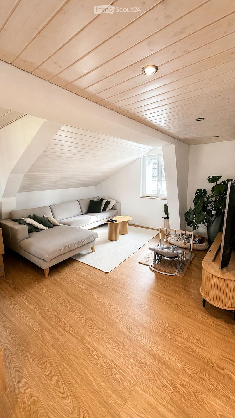 Modern living room with wooden floors, white walls, and a slanted wooden ceiling. Features a grey sectional sofa with pillows, a small coffee table, a round wooden table, a large plant, and a TV screen on a wooden stand.