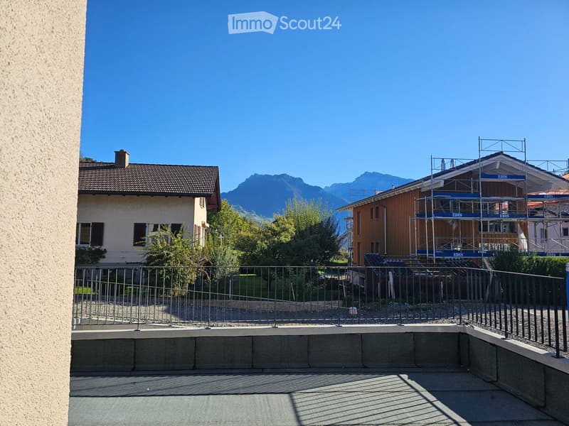 Balcony with metal railing, overlooking a residential area with houses, trees, and distant mountains.