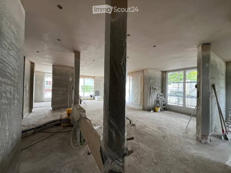Empty room with exposed concrete walls and floors, large windows, and construction materials scattered around.