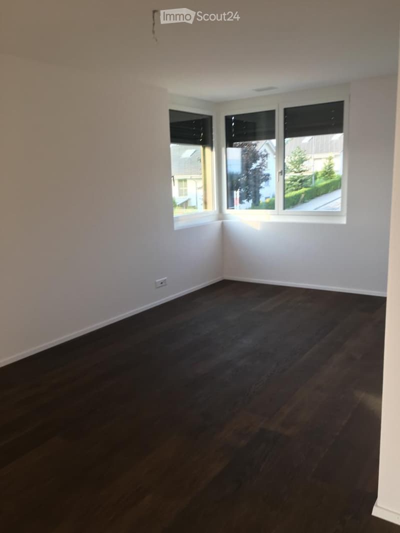 Empty room with dark wooden flooring and white walls, featuring three large windows with blinds and an electrical outlet on the wall.