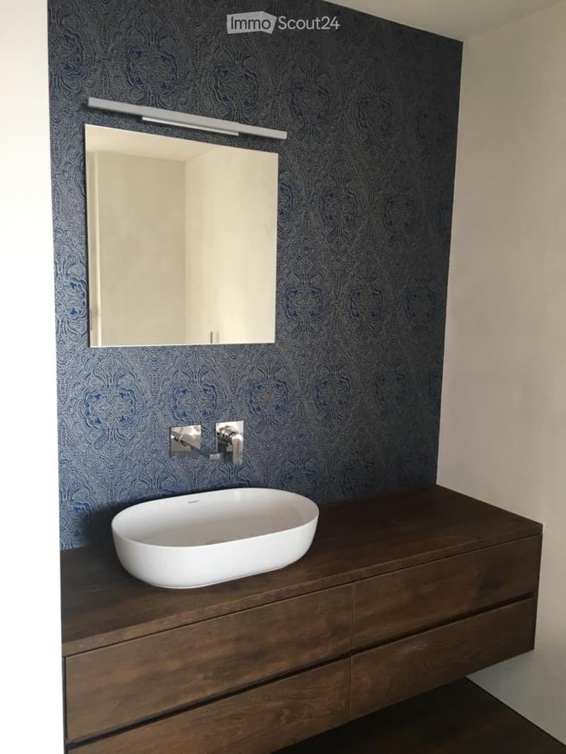 Modern bathroom with a dark blue patterned wall, a white sink on a dark wooden vanity, a large mirror with a built-in light fixture above it, and a sleek chrome faucet.