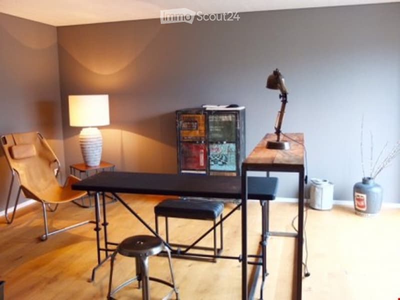 A modern, minimalist room with a wooden floor, grey walls, and a mix of contemporary and industrial furniture including a desk, chairs, and lamps.