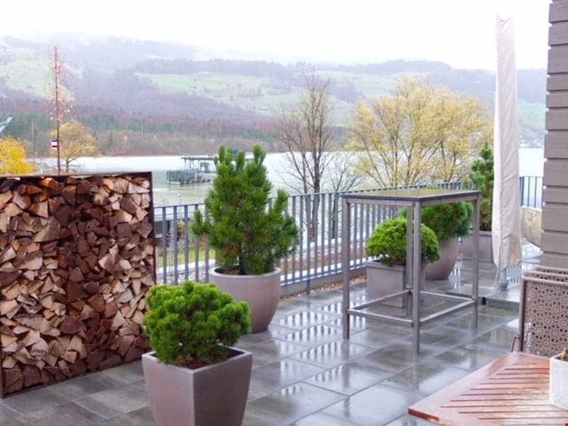 Balcony with a scenic view of a river and surrounding hills, featuring potted plants, a stack of firewood, and a metal table.