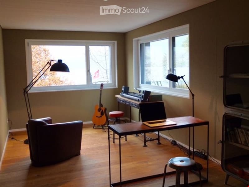A cozy, well-lit room with wooden flooring, large windows, a dark brown sofa, a wooden table with a laptop, a guitar, and a keyboard on a stand. Two floor lamps and two stools are also present.