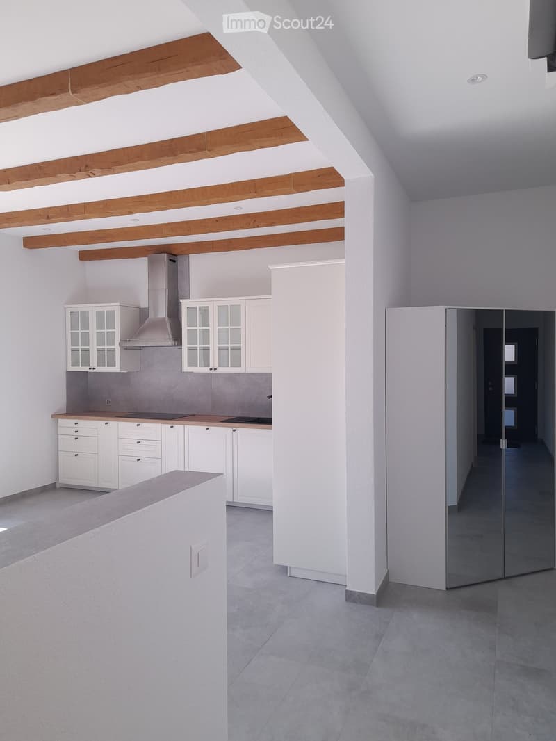 Modern kitchen with white cabinets, a range hood, and a central island. Exposed wooden beams on the ceiling and a grey tiled floor.